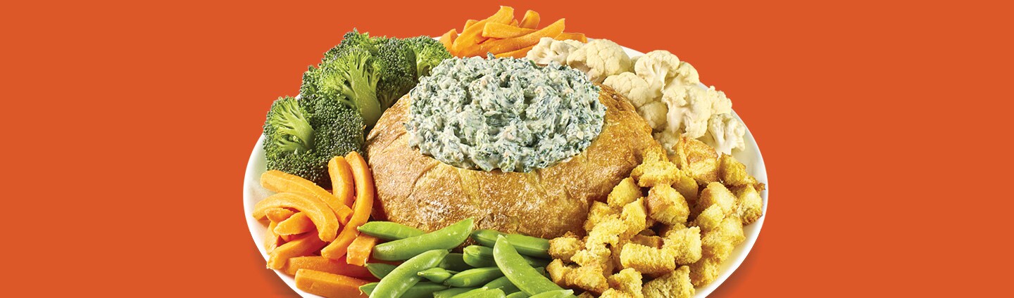 Bread bowl with spinach dip surrounded by veggies and bread cubes, on orange background