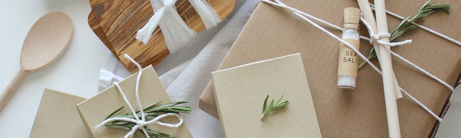 Wooden spoons, a vial of sea salt, cardboard, twine, rosemary and a cutting board wrapped in cheesecloth laid out on a white table